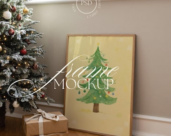 May include: A framed Christmas tree art print with a watercolor design. The artwork is displayed in a wooden frame, leaning against a wall. A decorated Christmas tree and wrapped gifts are in the foreground.