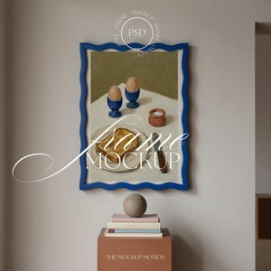 May include: A framed painting with a blue wavy frame depicts a breakfast scene with toast, eggs in blue egg cups, and a small pot. The words "Frame Mockup" are overlaid on the image. A sphere sits atop a stack of books.