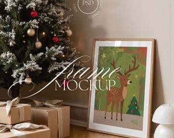 May include: A framed art print featuring a deer in a forest scene, leaning against a wall next to a decorated Christmas tree and wrapped gifts. The print has a wooden frame. The words "Frame Mockup" are visible on the image.