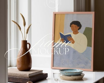 Op de afbeelding: Een ingelijste kunstdruk met een lichtroze lijst, met een persoon die een boek leest. Een bruine vaas met gedroogde planten, boeken en een klein bordje met een decoratief object staan op de voorgrond. De tekst "Frame Mockup" is over de afbeelding geplaatst.