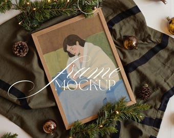 May include: A wooden framed art print featuring a woman in a white robe, with the text "Frame Mockup". The artwork is set on a green cloth with black stripes, decorated with pine branches, ornaments, and pinecones. The image is a mockup.