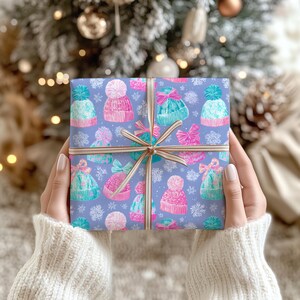 May include: A square gift box wrapped in light purple paper featuring a repeating pattern of colorful knit hats with pom-poms and bows, interspersed with white snowflakes. The package is tied with a thin, striped beige and white ribbon.