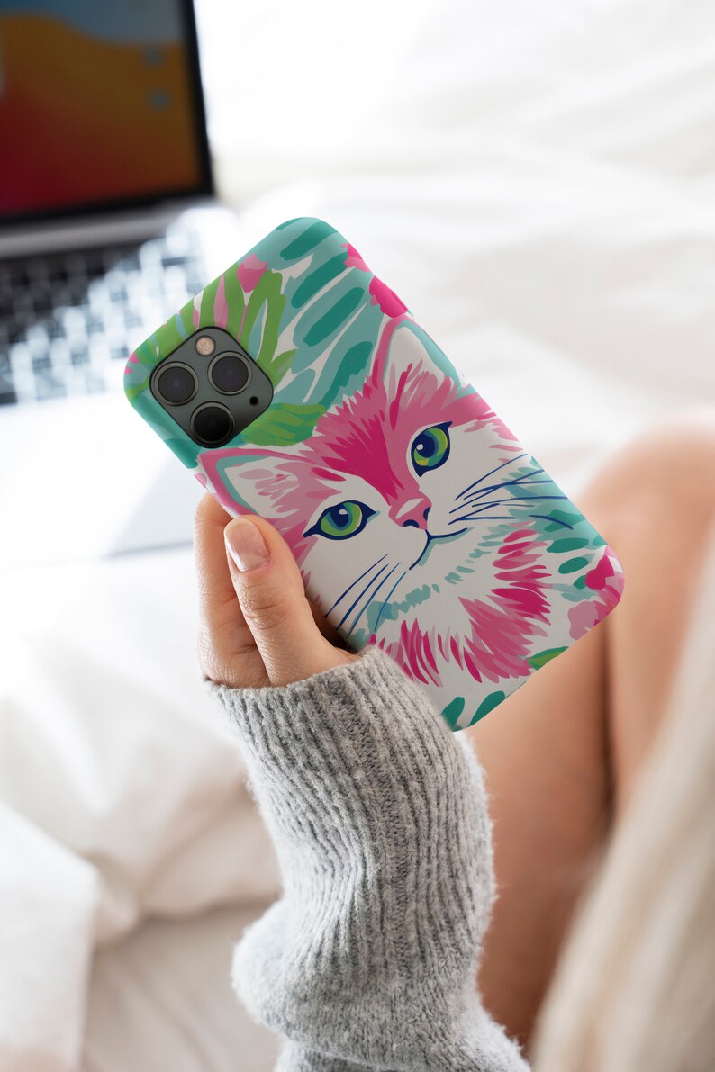 Pode incluir: Uma capa de telefone branca com uma ilustra&ccedil;&atilde;o de um gato rosa e verde e um padr&atilde;o de folhas tropicais.