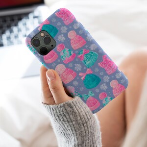 May include: A smartphone case with a light blue background featuring a repeating pattern of pink and teal knit beanies with pom-poms, interspersed with white snowflake illustrations. The case has a cutout for a triple-lens camera system.