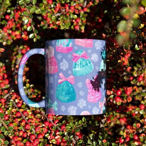May include: A ceramic mug with a light purple background featuring a repeating pattern of colorful knit winter hats in shades of pink and teal, accented with pink bows and white snowflake illustrations, complete with a matching purple handle.