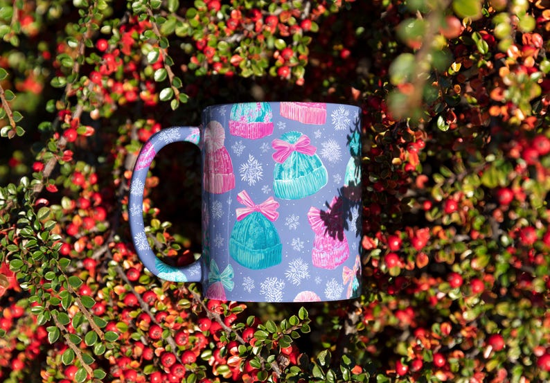 May include: A ceramic mug with a light purple background featuring a repeating pattern of colorful knit winter hats in shades of pink and teal, accented with pink bows and white snowflake illustrations, complete with a matching purple handle.