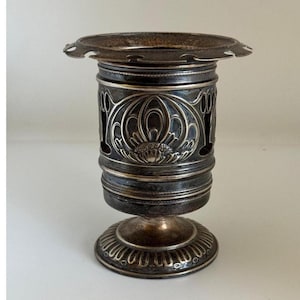 May include: A silver-plated, decorative, footed vase with an ornate design. The vase has a scalloped rim and a base with a floral pattern.