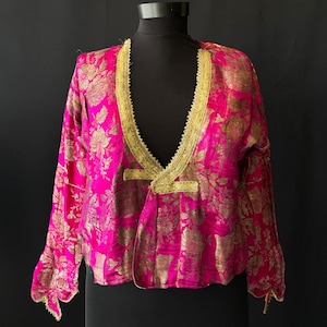 Pode incluir: Uma jaqueta fúcsia vibrante com um padrão de brocado floral em ouro metálico. A peça possui mangas compridas, um decote em V profundo e é finalizada com um acabamento trançado dourado decorativo ao longo da gola e fecho frontal.