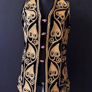 May include: Black velvet vest with gold embroidered leaf and swirl pattern. The V-neck vest is fastened with three pink buttons. The garment is displayed on a black mannequin.
