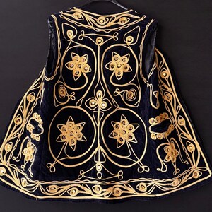 May include: A navy blue vest with intricate gold embroidery. The vest features floral and geometric patterns, with a detailed design around the edges. The garment is displayed on a black hanger against a dark background, highlighting the gold threadwork.