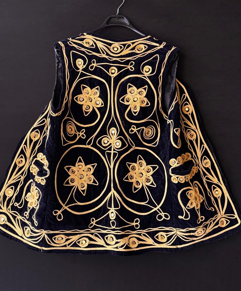 May include: A navy blue vest with intricate gold embroidery. The vest features floral and geometric patterns, with a detailed design around the edges. The garment is displayed on a black hanger against a dark background, highlighting the gold threadwork.
