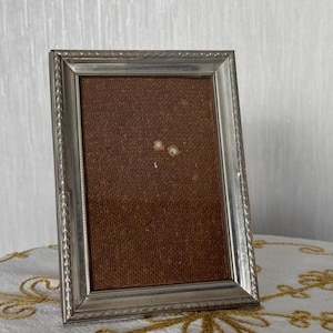 Puede incluir: Un marco de fotos plateado con un respaldo de tela marrón. El marco tiene un borde decorativo.