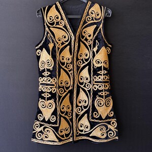 May include: Black velvet vest with gold embroidery featuring leaf and scroll patterns. The sleeveless vest has a V-neck and is suspended on a black hanger. The intricate gold embroidery stands out against the black fabric.