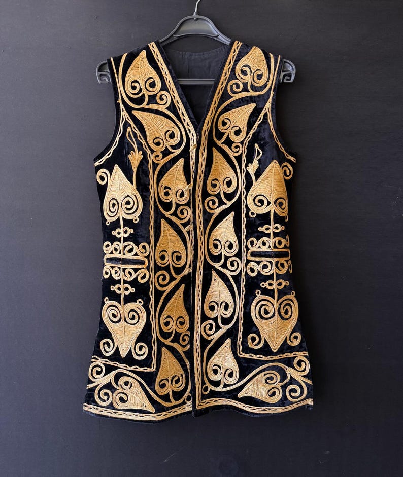 May include: Black velvet vest with gold embroidery featuring leaf and scroll patterns. The sleeveless vest has a V-neck and is suspended on a black hanger. The intricate gold embroidery stands out against the black fabric.