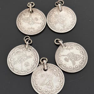 Puede incluir: Cinco colgantes de monedas antiguas de color plateado. Cada colgante redondo presenta un diseño ornamentado con un emblema central, un borde decorativo y pequeños detalles de estrellas. Los colgantes tienen un lazo en la parte superior para colgar.