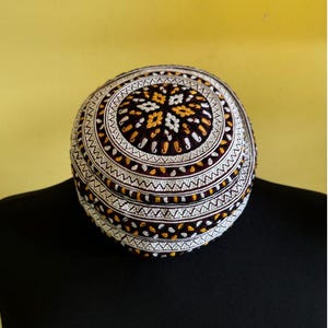 May include: A round, embroidered cap with a maroon base and white and gold embroidery. The cap has a geometric pattern with a central star.