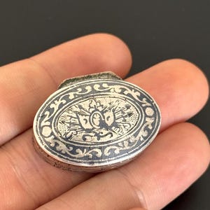 May include: An oval, silver-toned box with intricate engraved designs. The lid features a central oval with a detailed emblem, surrounded by ornate patterns. The box is held in a hand, showcasing its size and craftsmanship.
