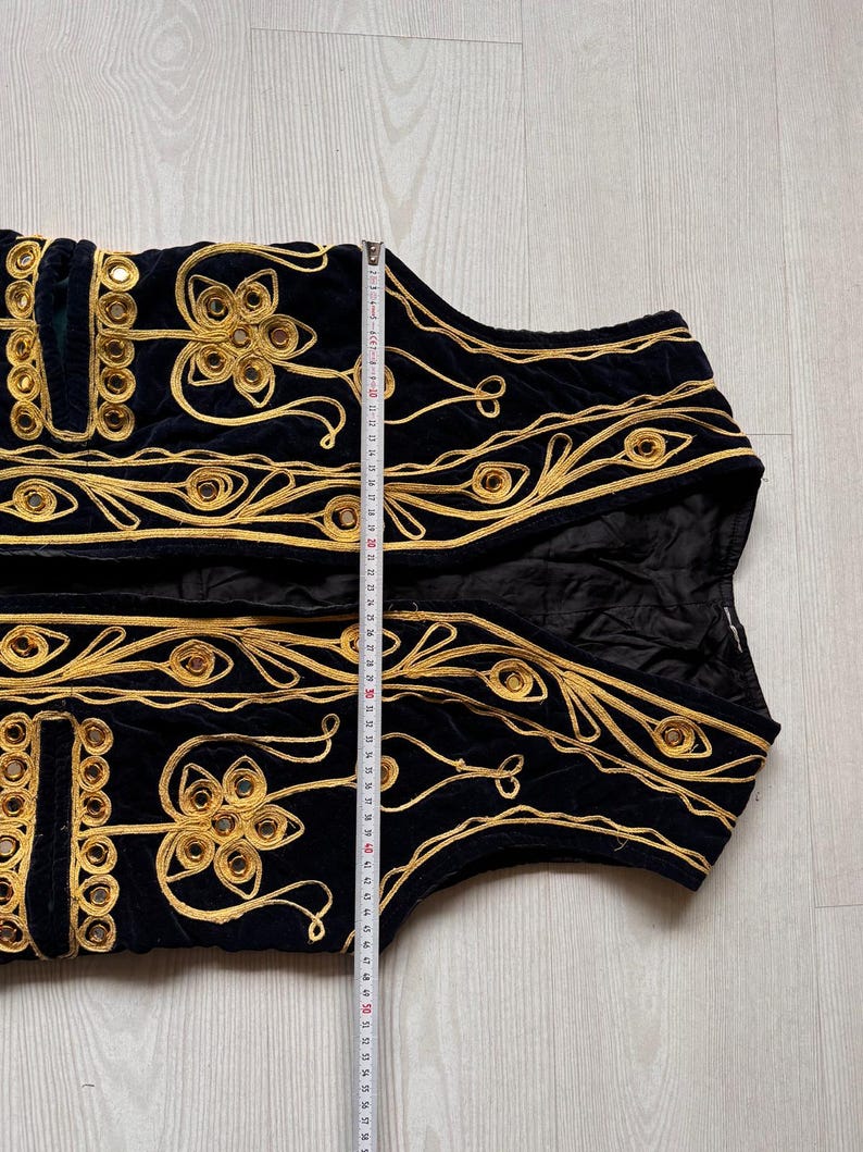 May include: Black velvet vest with gold embroidery and mirrored accents. The vest features intricate floral and geometric patterns. The length of the vest is approximately 20 inches.
