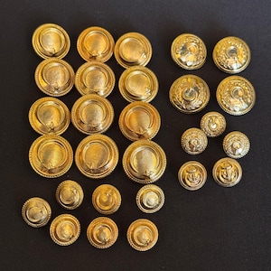 Puede incluir: Una colección de botones de metal en tono dorado de varios tamaños. La mayoría presenta un motivo de luna creciente y estrella con un acabado texturizado. Un grupo más pequeño a la derecha muestra patrones en relieve, incluyendo anclas.