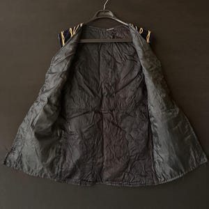 May include: A black vest with a quilted interior, hanging on a black hanger. The vest's exterior is navy blue with gold-colored trim along the shoulders. The vest is open, revealing the interior lining.
