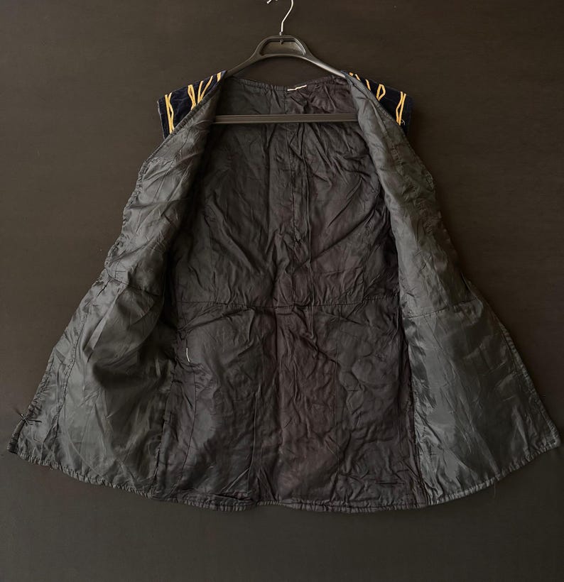 May include: A black vest with a quilted interior, hanging on a black hanger. The vest's exterior is navy blue with gold-colored trim along the shoulders. The vest is open, revealing the interior lining.