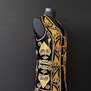 May include: Black velvet vest with gold embroidery. The vest features an intricate design of swirling patterns and leaf-like shapes. It has a V-neck and is fastened with small pink buttons. The vest is displayed on a black mannequin against a black background.