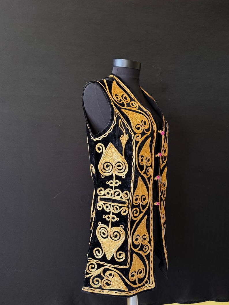 May include: Black velvet vest with gold embroidery. The vest features an intricate design of swirling patterns and leaf-like shapes. It has a V-neck and is fastened with small pink buttons. The vest is displayed on a black mannequin against a black background.