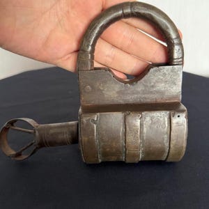 May include: A heavy, weathered iron padlock with a thick, rounded shackle and a cylindrical body. A long, metal key with a circular, open-work bow is inserted into the side of the lock, featuring a dark, oxidized patina and visible surface wear.