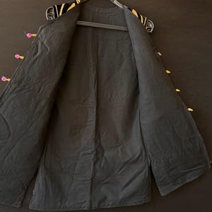 May include: Black jacket with gold and pink detailing. The jacket is open, showing the black lining. The collar and edges are decorated with gold embroidery. The jacket is hanging on a black hanger. The garment is a dark color.