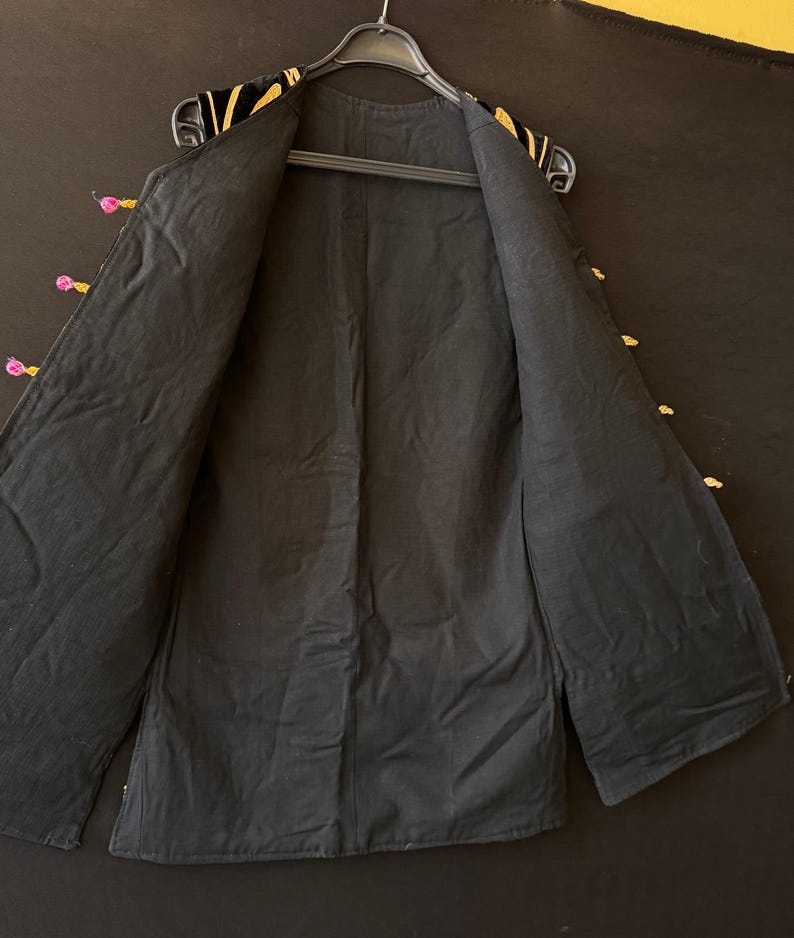 May include: Black jacket with gold and pink detailing. The jacket is open, showing the black lining. The collar and edges are decorated with gold embroidery. The jacket is hanging on a black hanger. The garment is a dark color.