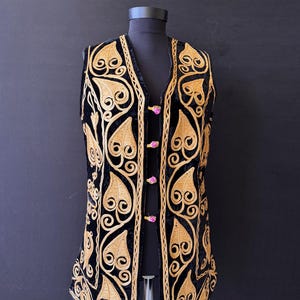 May include: A black velvet waistcoat featuring gold-coloured leaf and swirl embroidery. The waistcoat has a V-neck and is fastened with pink buttons. The embroidery is detailed and covers the front and edges, creating a striking contrast.