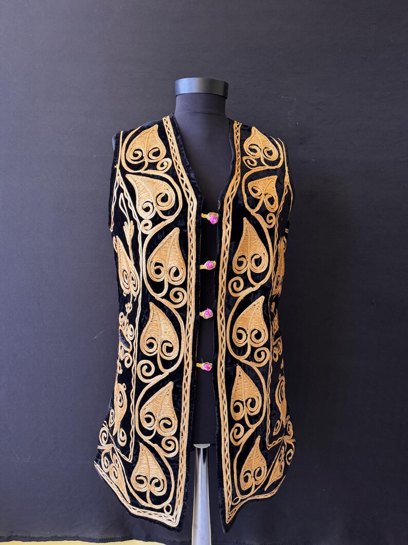 May include: A black velvet waistcoat featuring gold-coloured leaf and swirl embroidery. The waistcoat has a V-neck and is fastened with pink buttons. The embroidery is detailed and covers the front and edges, creating a striking contrast.