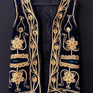 May include: A navy blue velvet vest with elaborate gold embroidery and mirrored accents. The vest features a floral and geometric design, with a teal green pocket detail. The vest is sleeveless and hangs on a black hanger against a dark background.