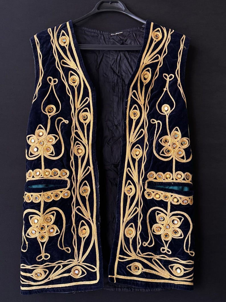 May include: A navy blue velvet vest with elaborate gold embroidery and mirrored accents. The vest features a floral and geometric design, with a teal green pocket detail. The vest is sleeveless and hangs on a black hanger against a dark background.