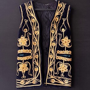 May include: A navy blue velvet vest with elaborate gold embroidery and mirrored accents. The sleeveless garment features a V-neck and is suspended from a black hanger against a dark background. The embroidery forms floral and geometric patterns.