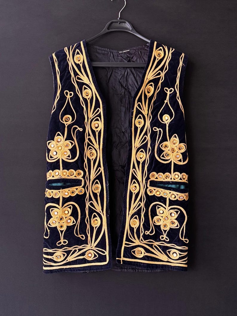 May include: A navy blue velvet vest with elaborate gold embroidery and mirrored accents. The sleeveless garment features a V-neck and is suspended from a black hanger against a dark background. The embroidery forms floral and geometric patterns.