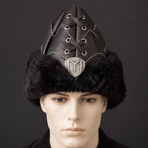 Puede incluir: Un sombrero de cuero negro con corona puntiaguda que presenta un diseño de cordones con ojales de metal plateado y costuras blancas. La base del sombrero está adornada con piel sintética negra gruesa y un emblema de metal plateado central.