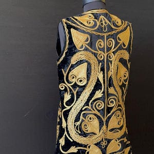 May include: Black velvet vest with gold embroidery. The sleeveless vest has a detailed floral and leaf pattern, creating a luxurious design. The vest is displayed on a mannequin against a dark background.