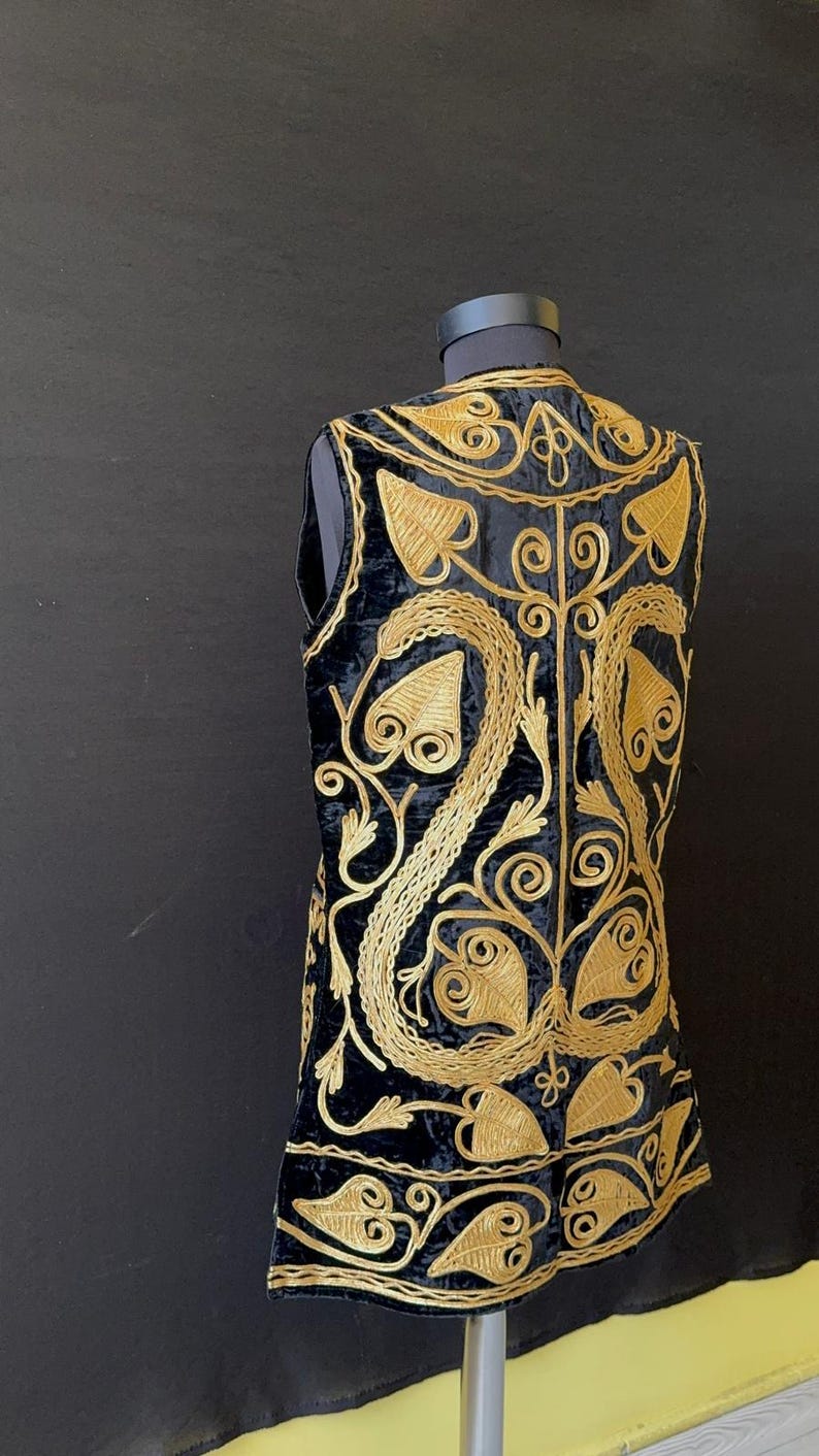 May include: Black velvet vest with gold embroidery. The sleeveless vest has a detailed floral and leaf pattern, creating a luxurious design. The vest is displayed on a mannequin against a dark background.