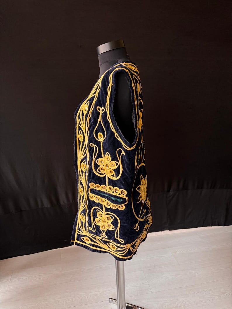 May include: A navy blue vest with intricate gold embroidery. The vest features a V-neck and a sleeveless design. The gold embroidery creates floral and geometric patterns across the front and sides of the vest. The vest is displayed on a mannequin.