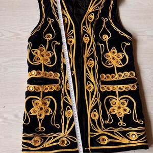 May include: Black velvet vest with gold embroidery and decorative mirrored accents. The vest features a V-neck and two front pockets. The gold embroidery includes floral and scroll designs. A measuring tape is placed along the vest, indicating its length.