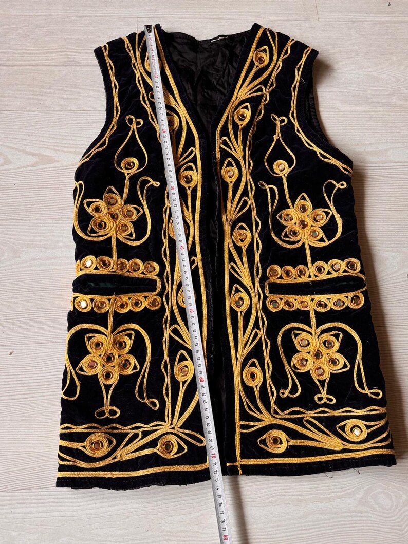 May include: Black velvet vest with gold embroidery and decorative mirrored accents. The vest features a V-neck and two front pockets. The gold embroidery includes floral and scroll designs. A measuring tape is placed along the vest, indicating its length.