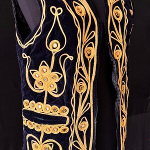 May include: Navy blue velvet vest with gold embroidery and mirror accents. The vest features intricate floral and geometric patterns. The gold embroidery outlines the edges and designs, creating a striking contrast against the dark blue fabric.