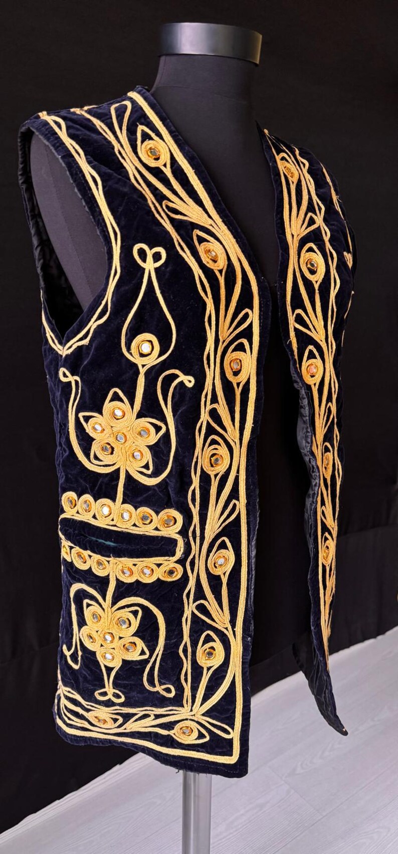 May include: Navy blue velvet vest with gold embroidery and mirror accents. The vest features intricate floral and geometric patterns. The gold embroidery outlines the edges and designs, creating a striking contrast against the dark blue fabric.