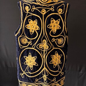 May include: A navy blue vest with intricate gold embroidery. The vest features a quilted design with floral and geometric patterns. The gold embroidery creates a striking contrast against the dark blue fabric, adding a touch of elegance and sophistication.