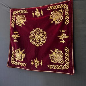 Può includere: Un tessuto quadrato in velluto bordeaux con ricami dorati intricati. Il design presenta motivi floreali e geometrici, tra cui un medaglione centrale e dettagli agli angoli. I bordi sono rifiniti con un bordo dorato.
