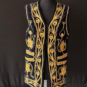 May include: A navy blue velvet vest with elaborate gold embroidery. The vest features a V-neck, two pockets, and a detailed floral pattern. The gold embroidery is intricately designed, creating a striking contrast against the dark blue fabric. The vest is displayed on a mannequin.
