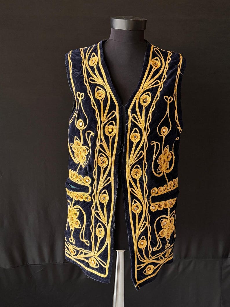 May include: A navy blue velvet vest with elaborate gold embroidery. The vest features a V-neck, two pockets, and a detailed floral pattern. The gold embroidery is intricately designed, creating a striking contrast against the dark blue fabric. The vest is displayed on a mannequin.