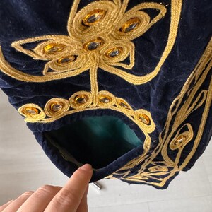 May include: Close-up of a navy blue velvet garment with intricate gold embroidery. The embroidery features floral and geometric patterns, embellished with amber-colored jewels. A pocket with a green lining is visible.