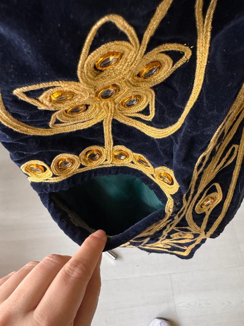 May include: Close-up of a navy blue velvet garment with intricate gold embroidery. The embroidery features floral and geometric patterns, embellished with amber-colored jewels. A pocket with a green lining is visible.